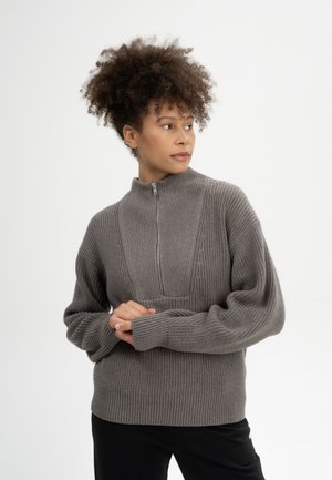 Jonge vrouw met krullend haar die een grijze gebreide trui met rits draagt, kijkt naar haar rechterkant met handen voor zich gevouwen tegen een effen achtergrond.