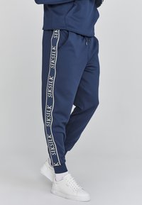 Marineblauwe sweatpants van een gestructureerde stof, met witte "SIKSILK" logo-stripes langs de zijkanten, een elastische tailleband en boorden.