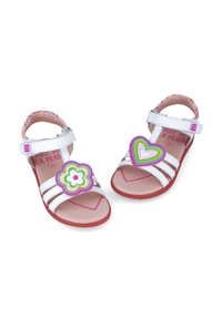 Sandalias blancas con diseño de tres correas, acentos en rosa, adornadas con una flor multicolor y un corazón en la parte frontal. Suela de goma con contorno.