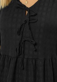 Top negro texturizado con escote en V, que presenta un detalle de lazo con cordón y un dobladillo acampanado. La tela tiene un sutil patrón de cuadrícula.