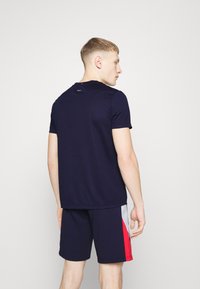 Camiseta deportiva de color navy con mangas cortas y escote redondo, combinada con pantalones cortos navy que tienen acentos laterales en rojo y gris.