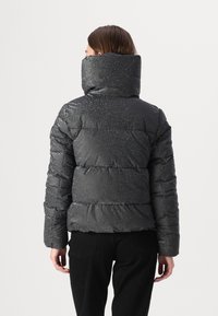 Zwarte pufferjack met hoge kraag, met een glanzend, gestructureerd oppervlak en een doorgestikte uitstraling. Gezien vanuit de achterkant, gecombineerd met zwarte broeken.