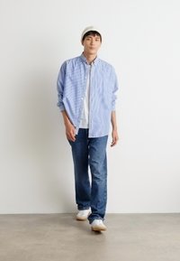 Gestreiftes blaues Hemd über einem weißen T-Shirt, kombiniert mit blauen Jeans und weißen Sneakern. Modell, das vor einem neutralen Hintergrund läuft.