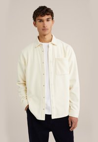 Cream-colored fleece shirt jacket with a relaxed fit, front pocket, and snap-button closure, worn over a white crewneck T-shirt.
