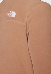 Giacca in pile marrone con una texture morbida, dotata di un logo ricamato bianco sulla spalla sinistra e cuciture a contrasto.