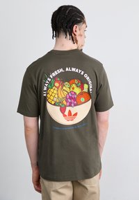 Olivenfarvet bomulds t-shirt med et farverigt frugtskål grafik på ryggen, med tekst og Adidas-logo i røde og blå accenter.