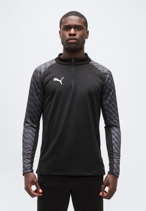 Νεαρός άνδρας που φοράει μαύρο αθλητικό τοπ Puma με μακριά μανίκια και σχέδιο στα μανίκια, στέκεται μπροστά σε απλό φωτεινό φόντο.