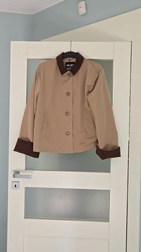 Bežna jakna s rjavim ovratnikom in manšetami, z zapiranjem na gumb in dvema stranskima žepoma, obešena na bež vratih. Material je videti gladek.