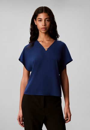 Jeune femme aux cheveux ondulés foncés, portant un chemisier bleu à col en V et manches courtes et un pantalon noir, sur un fond gris uni.