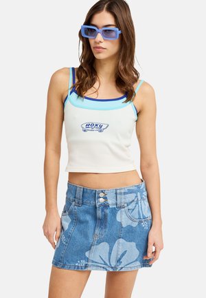 Młoda kobieta w niebiesko przyciemnianych okularach przeciwsłonecznych, białym krótkim topie z niebieskim wykończeniem i logo deskorolki oraz dżinsowej spódnicy z dużymi kwiatowymi wzorami.