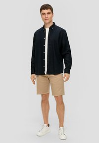 Donkerblauwe overhemdbloes over een wit T-shirt, gecombineerd met beige shorts en witte sneakers. Model staat voor een neutrale achtergrond.