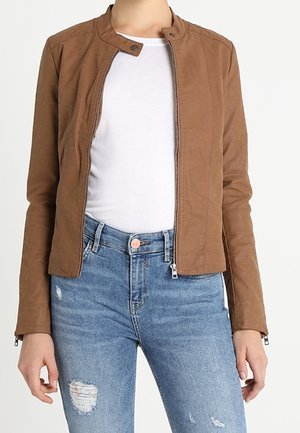 Person iført en brun lynlåstrøje over en hvid skjorte kombineret med revnede blå jeans, stående mod en ensfarvet lys baggrund.