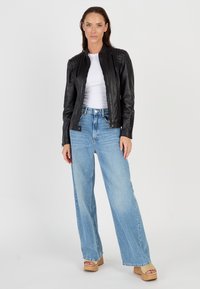 Zwarte leren jack met gestructureerde schouders, gedragen over een witte top. Gecombineerd met lichte blauwe wijde jeans en tan platform sandalen.