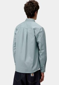 Camisa de botones azul claro con detalle de pliegue en la parte posterior, mangas largas y puños abotonados. Fabricada en una tela suave. Usada con pantalones de mezclilla oscuros.