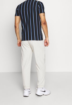Mand stående med ryggen mod kamera iført sort T-shirt med blå striber, hvide joggingbukser og hvide Nike sneakers på en ensfarvet baggrund.