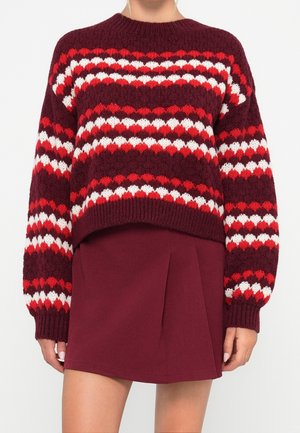 Femme portant un pull en tricot à motifs bordeaux, rouge et blanc avec des manches longues et une jupe plissée bordeaux.