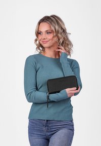 Black zippered wallet with a textured finish, held against a teal ribbed sweater. Features a minimalist logo on the front.