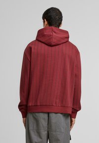 Sweat-shirt à capuche bordeaux avec un motif à rayures verticales, des poignets et un ourlet côtelés. Tissu doux, coupe ample, présentant une couture centrale au dos.