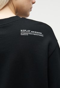 Άτομο με κοντά ξανθά μαλλιά που φοράει μαύρο πουλόβερ με λευκή επιγραφή "REPLAY ICON SELECTION NOT ORDINARY PEOPLE Registered Trade Mark" στην πλάτη.