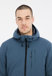 Giacca blu con cappuccio, dotata di zip frontale, cappuccio regolabile con cordino e tasca laterale. Realizzata in materiale liscio e resistente all'acqua.