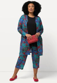 Ensemble à imprimé floral multicolore composé d'un long cardigan ouvert et de pantalons cropped, en tissu léger. Accessoirisé avec une pochette matelassée rouge.
