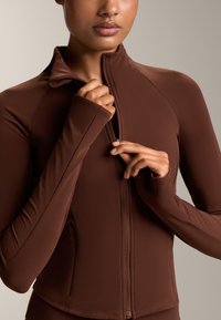 Smešani smeđi sportski zip-up jakna sa uskim krojem, visokim ovratnikom, dugim rukavima i detaljima s patent zatvaračem. Mekan, rastezljiv materijal s minimalnim šavovima.