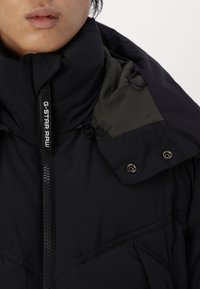 Melna polsterēta jaka ar augstu apkakli, kurai ir tumši zaļa iekšējā oderējuma un priekšējais rāvējslēdzis. Ievērojama aparatūra ietver spiedpoges.