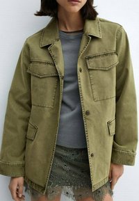 Žena v olivově zelené bundě s kapsami na klopě přes šedou košili a zelenou květinovou krajkovou sukni.