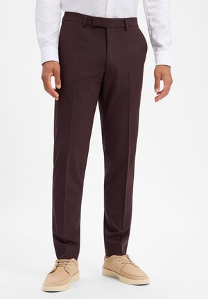 Mann trägt dunkelbraune Anzughose, weißes langärmeliges Hemd und hellbeige Freizeitschuhe, steht vor einem einfarbigen weißen Hintergrund.