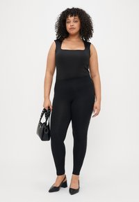 Μαύρο κορμάκι με τετράγωνη λαιμοκοψιά, σε συνδυασμό με μαύρα κολάν που έχουν λεπτομέρεια στο πλάι; το μοντέλο κρατάει μια μικρή μαύρη τσάντα και φοράει μαύρες γόβες.