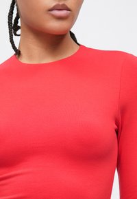 Nærbilde av en person med flettet hår som har på seg en ensfarget rød genser med lange ermer foran en hvit bakgrunn.