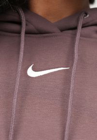 Barna Nike kapucnis pulóver fehér logóval, mely zsinóros kapucnit, texturált anyagot és varrott varratokat tartalmaz. Közeli kép a nyakkivágásról.