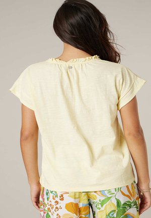 Vrouw met lang donker haar draagt een lichtgeel kortärmelig shirt met een gerimpelde halslijn en een broek met bloemenpatroon, gezien van achteren.