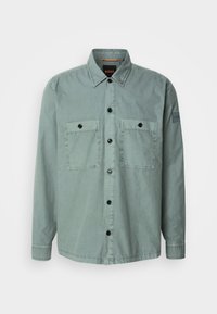 Camisa de botones verde claro, hecha de una tela suave, con dos bolsillos en el pecho y botones negros. Cuello clásico y mangas largas con puños abotonados.