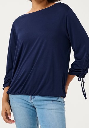 Vrouw draagt een marineblauwe top met lange mouwen met geplooide trekkoorden aan de mouwen en lichtblauwe spijkerbroek, staat met hand op heup.