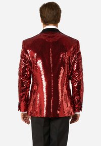 Sarkans sekmēts bleizeris ar spīdīgu apdari, ar melnu apkakli un garām piedurknēm, uzvilkts virs melniem biksēm. Rādīts aizmugures skats.