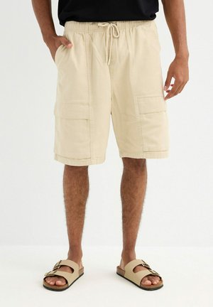 Hombre con pantalones cortos de cargo beige hasta la rodilla con cordón y sandalias marrón claro con hebilla sobre un fondo blanco.