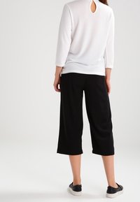 Haut blanc à manches longues avec une découpe en forme de trou de serrure dans le dos, associé à un pantalon noir ample et court, et des chaussures noires à enfiler.