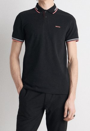 Mann trägt schwarzes Poloshirt mit rot-weiß gestreiftem Kragen und Ärmelabschluss, Logo "BOSS" auf der Brust, Hände in den Taschen.