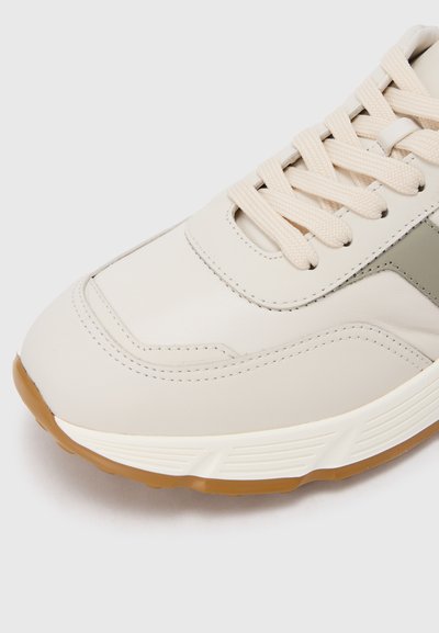 Baskets beige avec une tige en cuir lisse, une semelle en caoutchouc texturée et des accents gris contrastants. Dispose de lacets plats et de détails cousus.