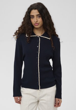 Jonge vrouw met krullend haar draagt een marineblauwe geribbelde cardigan met knopen en crèmekleurige bies en witte broek, staand tegen een effen achtergrond.