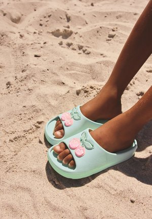 Pieds portant des sandales bleu clair à bout ouvert avec des décorations cerises roses, debout sur une plage de sable clair.