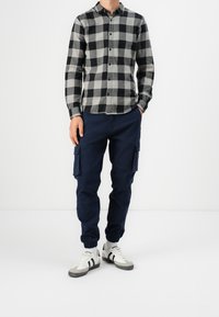 Camisa de franela a cuadros en negro y gris, pantalones cargo navy con bolsillos laterales, y zapatillas blancas con rayas negras. Atuendo casual.