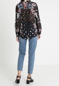 Gennemsigtig sort bluse med blomstermønster på skuldrene, prikker på ryggen og lange ærmer. Kombineret med lyseblå jeans og sorte sandaler.