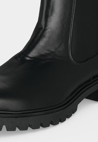 Gros plan d’une botte chevillée en cuir noir avec un panneau latéral élastique et une semelle épaisse à rainures.