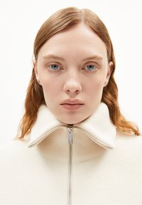 Beige high-neck sweater with a full zipper, made from textured knit fabric. Features a prominent ribbed collar and subtle stitching details.