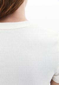 Bílý hladký top s krátkým rukávem, který se vyznačuje jemnou texturou a kulatým výstřihem. Zadní pohled ukazuje klesající linie od ramene k lemu.