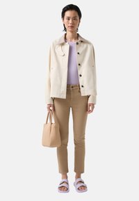 Light beige jacket with a textured collar, purple top, tan fitted pants, and lavender sandals. Holds a small beige handbag with shoulder straps.