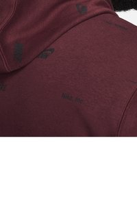 Burgundy hoodie made of soft fabric, featuring a textured surface with black Nike logos and branding subtly printed on the material.