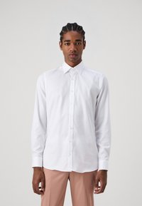 Chemise blanche à manches longues avec un col classique et des boutons sur le devant. Tissu lisse, coupe ajustée et design épuré. Associée à un pantalon léger rose.